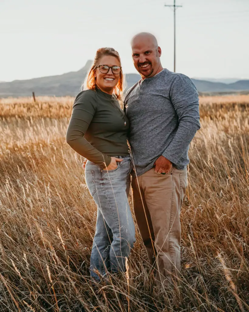 Smiling couple, founders of TH Custom Builder, standing in a golden Wyoming field at sunset with mountains in the background